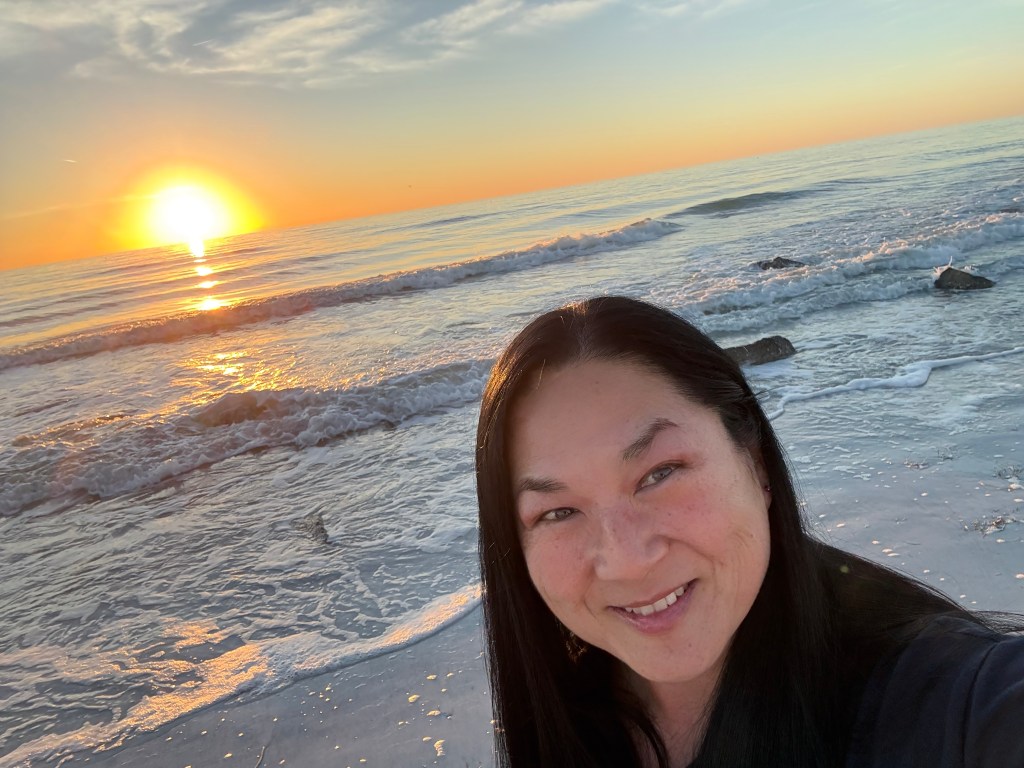 A woman with long black hair smiles at the camera, standing on a beach at sunrise or sunset. The sun is setting over the o...