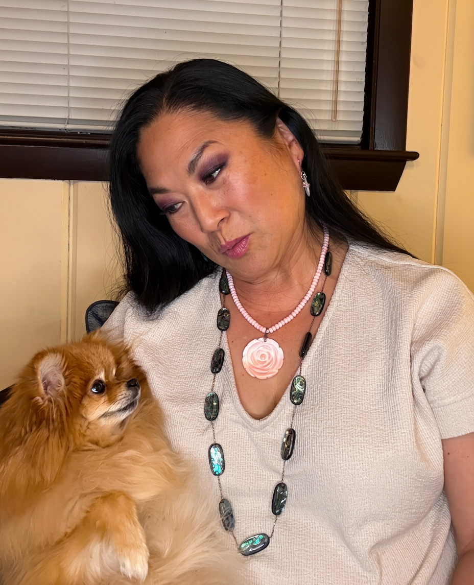 A woman with long black hair, wearing a white shirt and a necklace, sits in front of a window with a small dog on her lap....