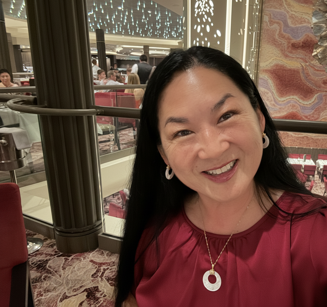 Subtle professional portrait retouch of a smiling woman seated at a restaurant table on a cruise ship. Maintain natural skin texture and authenticity. Slightly brighten overall exposure, gently soften under-eye shadows, even out skin tone without over-smoothing, enhance eye clarity slightly, and apply very light color correction for warm, balanced tones. Keep it realistic and not over-processed.